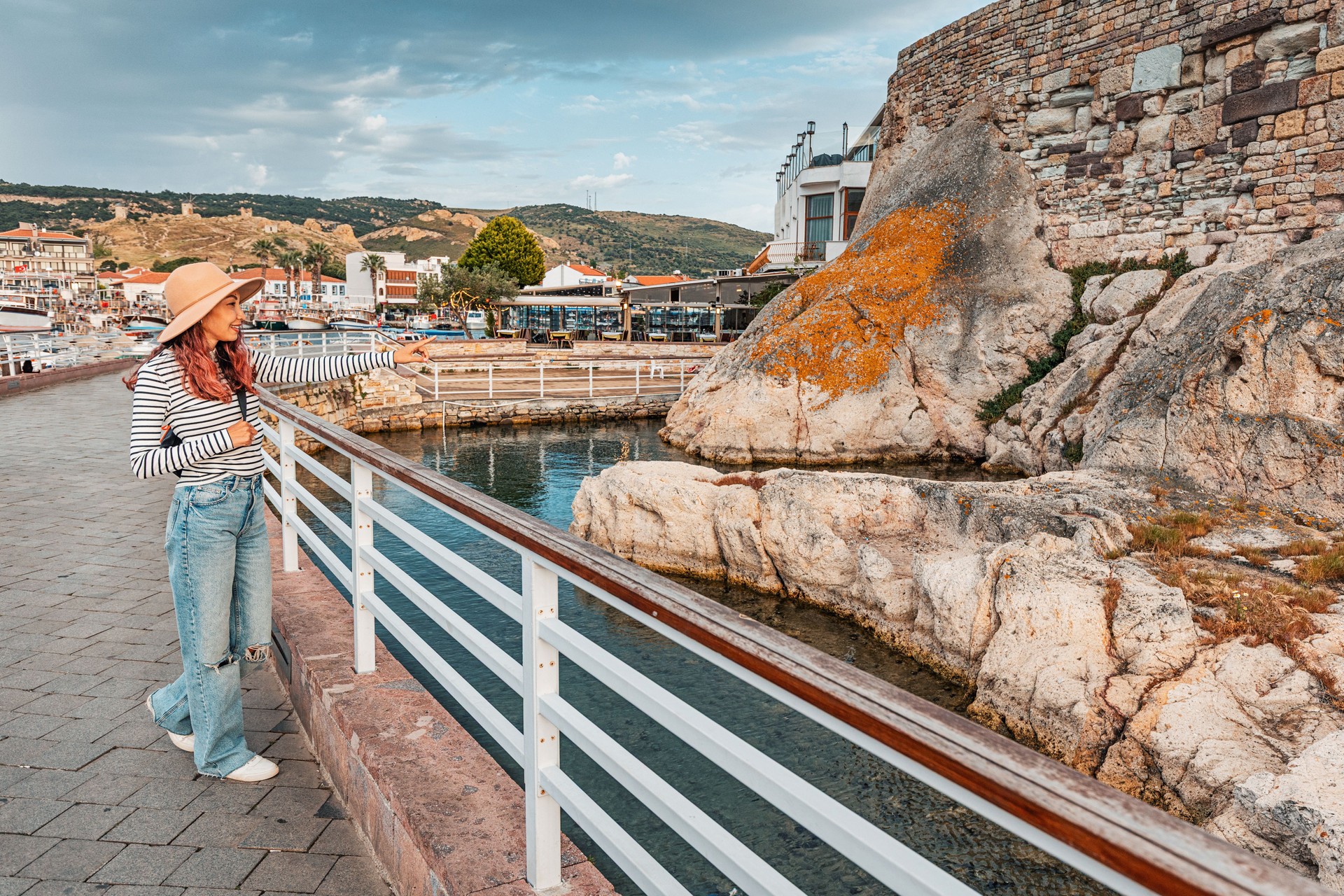 Woman in a hat admiring ancient stone castle walls by the sea in Foca, Turkiye, with a scenic mountainous backdrop